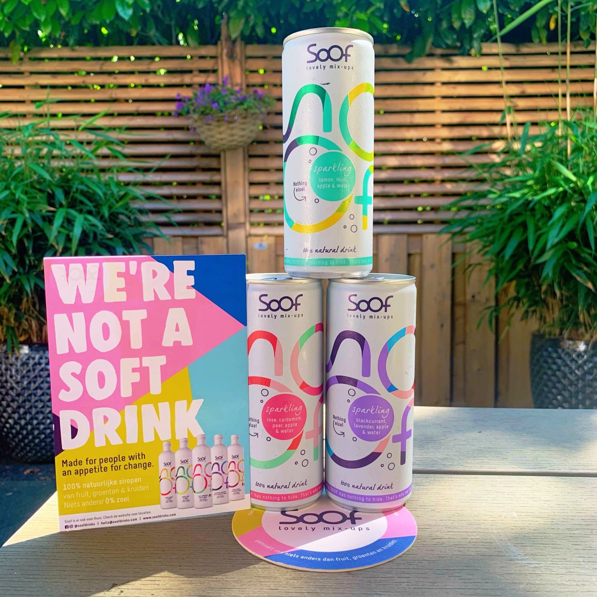 Een display met blikjes Soof-drank en promotiemateriaal op een terras. Inclusief een bord, onderzetters en gestapelde blikjes.