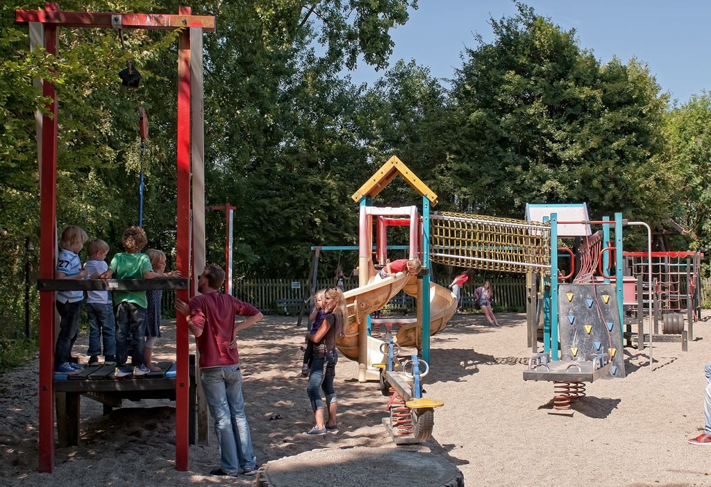 Kinderen spelen op speeltoestellen: schommels, een klimmuur en platforms. Bruine grindgrond en bomen.