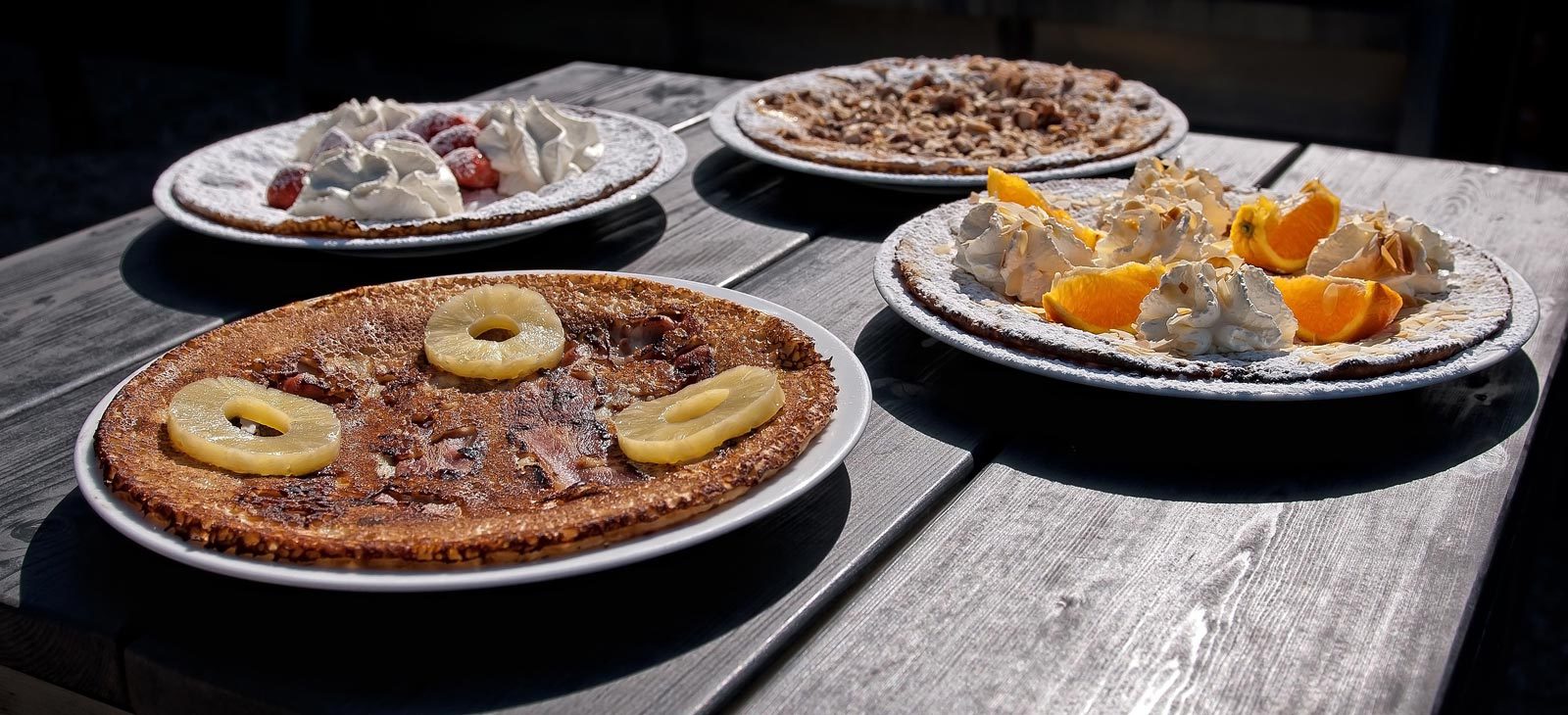 Vier borden met pannenkoeken, belegd met diverse soorten fruit en slagroom, staan op een houten tafel.