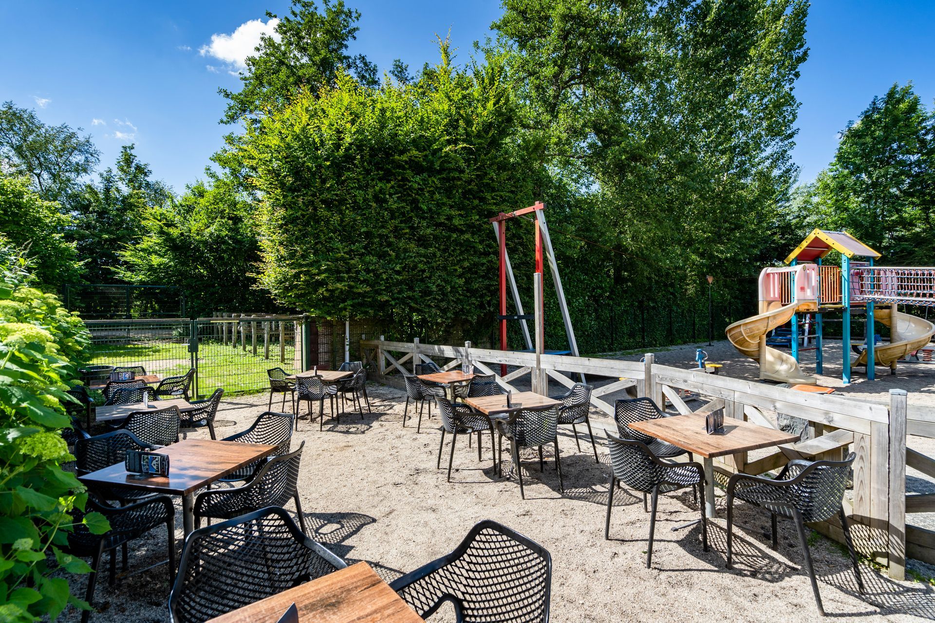 Buitenrestaurant met tafels en stoelen op het zand, naast een speeltuin en groene bomen.