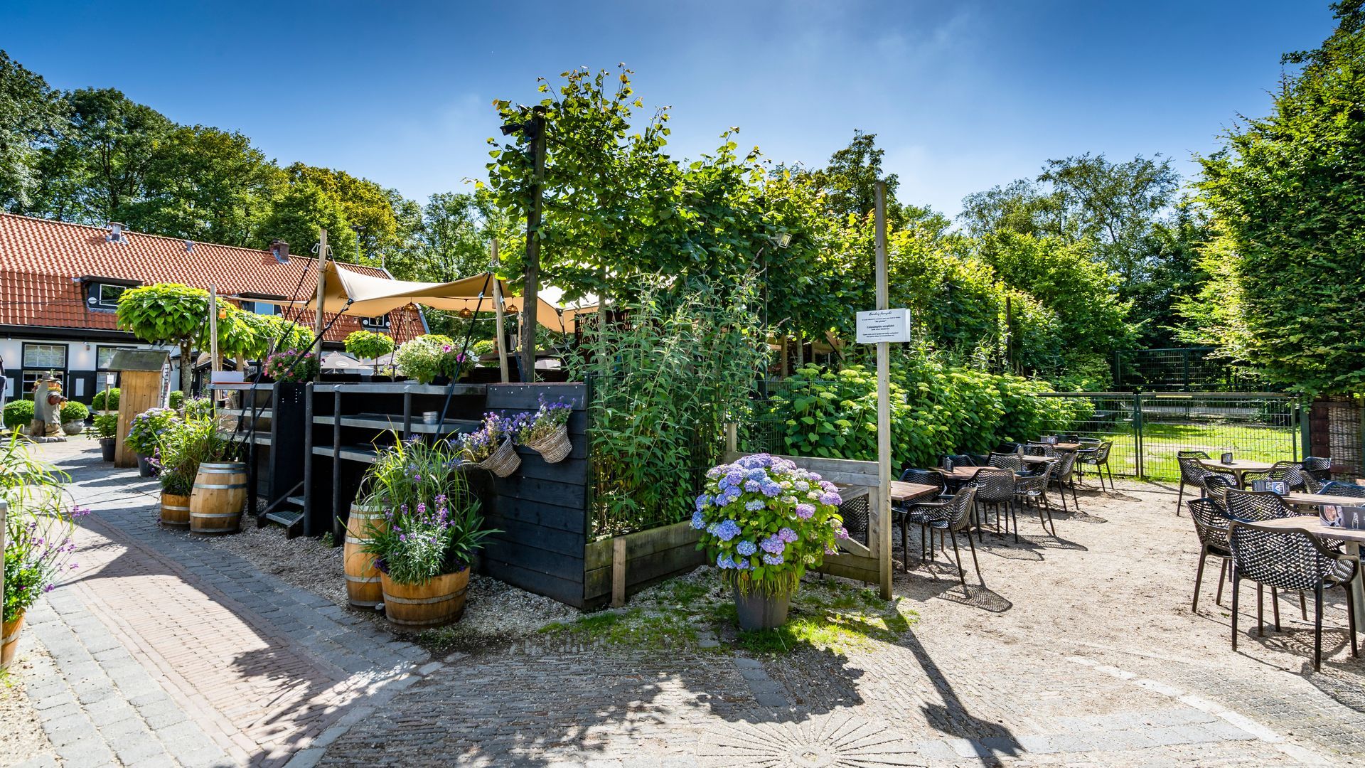 Een terrascafé met tafels, planten en een vakwerkgebouw onder een zonnige hemel.