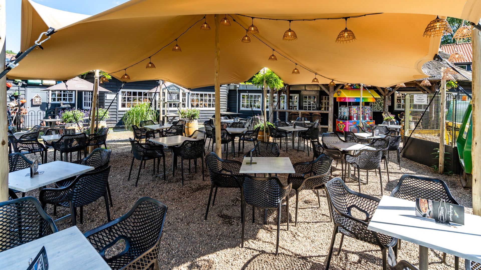 Terrascafé met zwarte stoelen en tafels onder een beige luifel. Boven de luifel hangen lichtslingers.