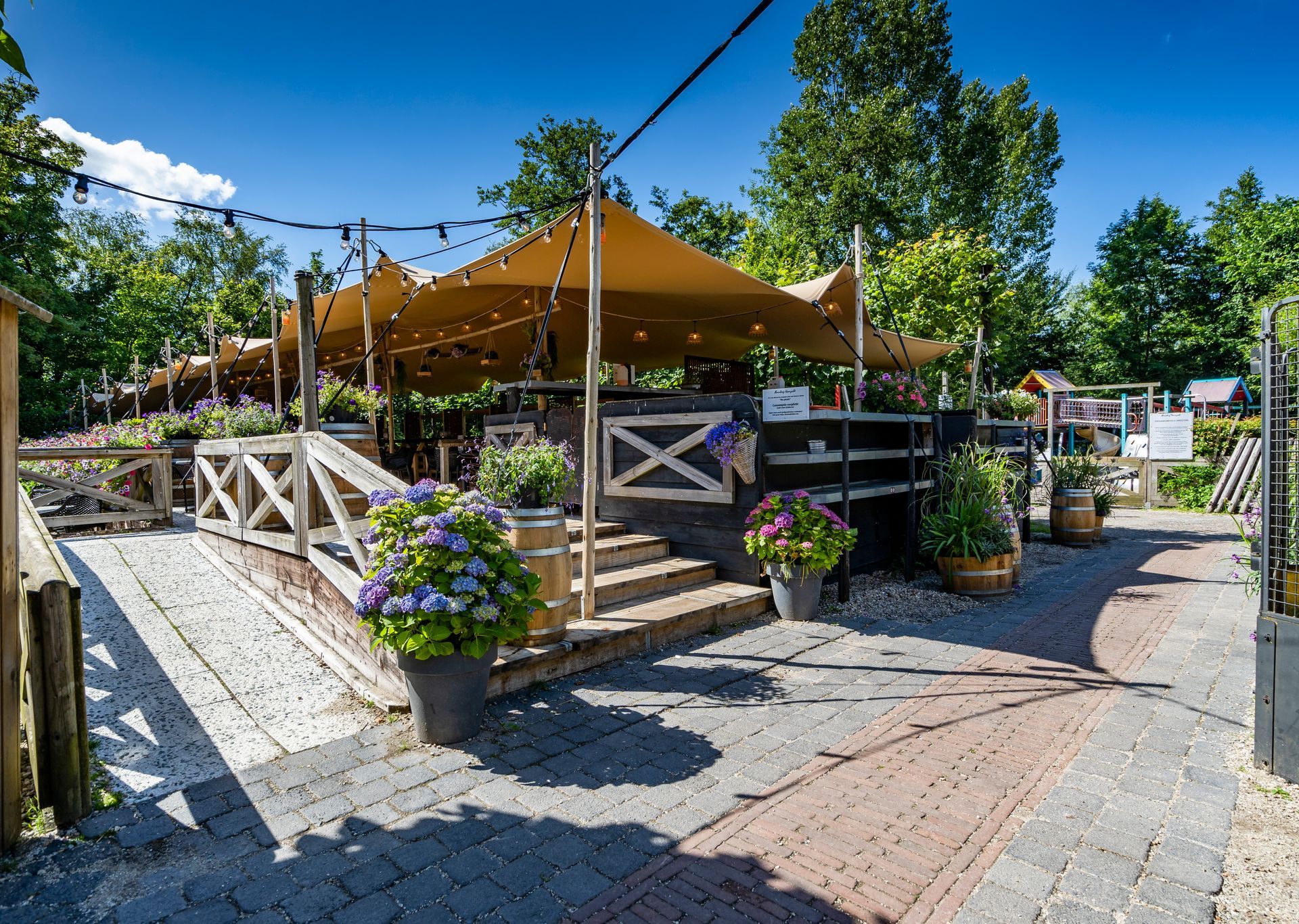 Buitenbar met beige luifels, houten balustrade, bakstenen pad, potplanten en lichtslingers.