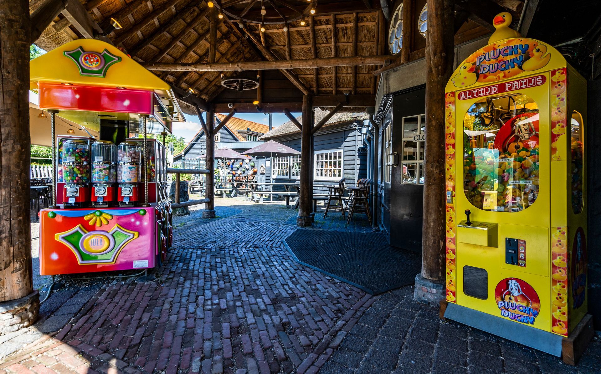 Twee kleurrijke speelautomaten onder een houten afdak; een bakstenen pad leidt naar een buitenruimte.