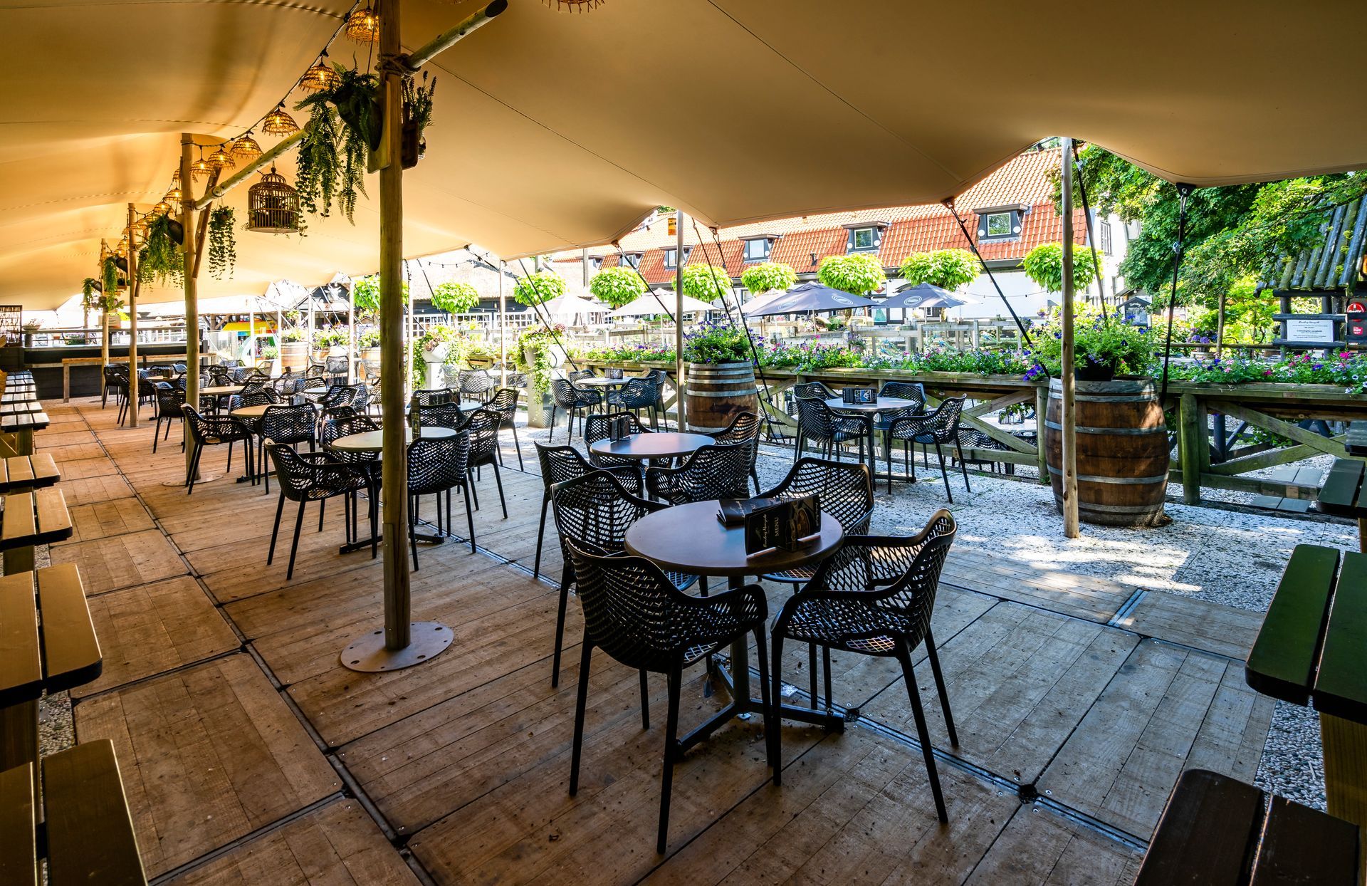Terras met zitplaatsen onder een beige luifel. Tafels en stoelen staan opgesteld op een houten vlonder. Gebouwen en bomen op de achtergrond.