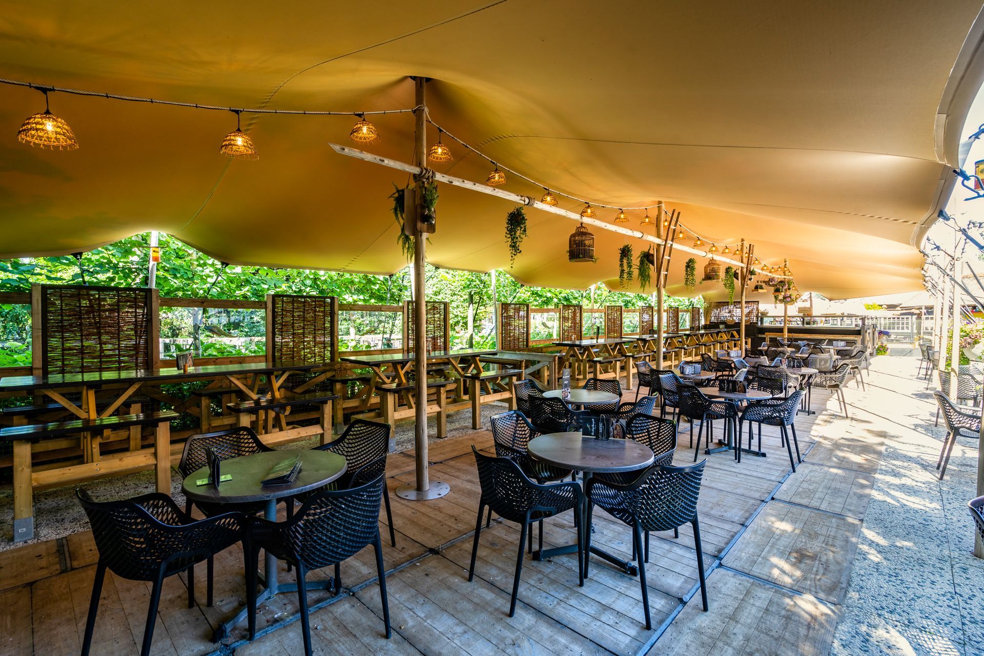 Een terrascafé met tafels, zwarte stoelen en een canvas dak, onder de bomen.