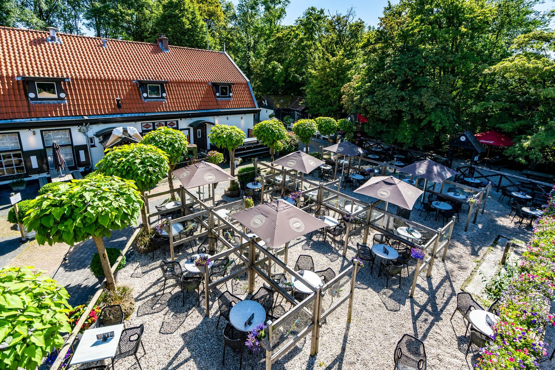Een restaurant in de openlucht met tafels, parasols en groene bomen rondom een ​​bakstenen gebouw.