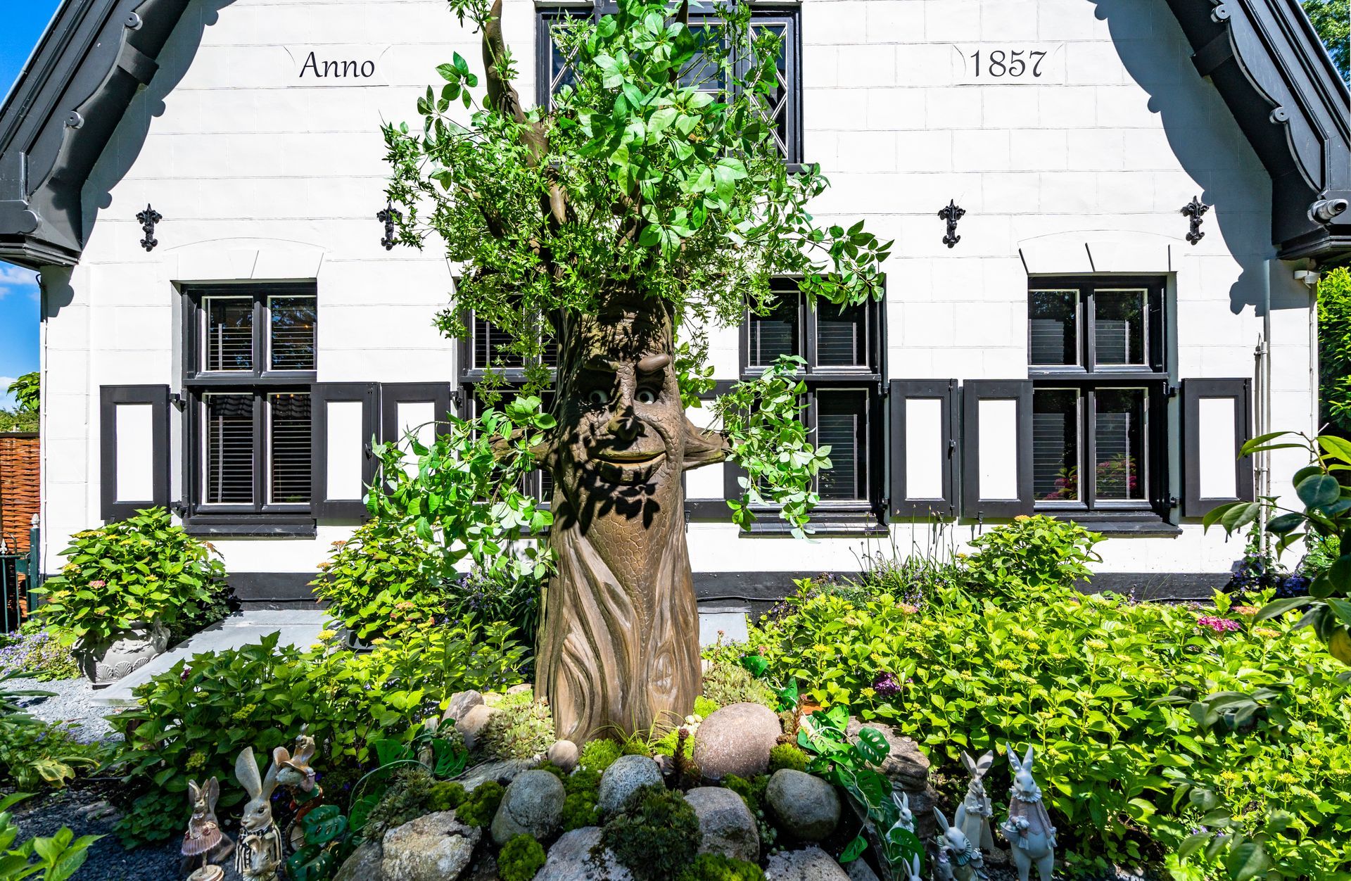 Wit huis met zwarte kozijnen en luiken, voor een stenen sculptuur en weelderige groene struiken.