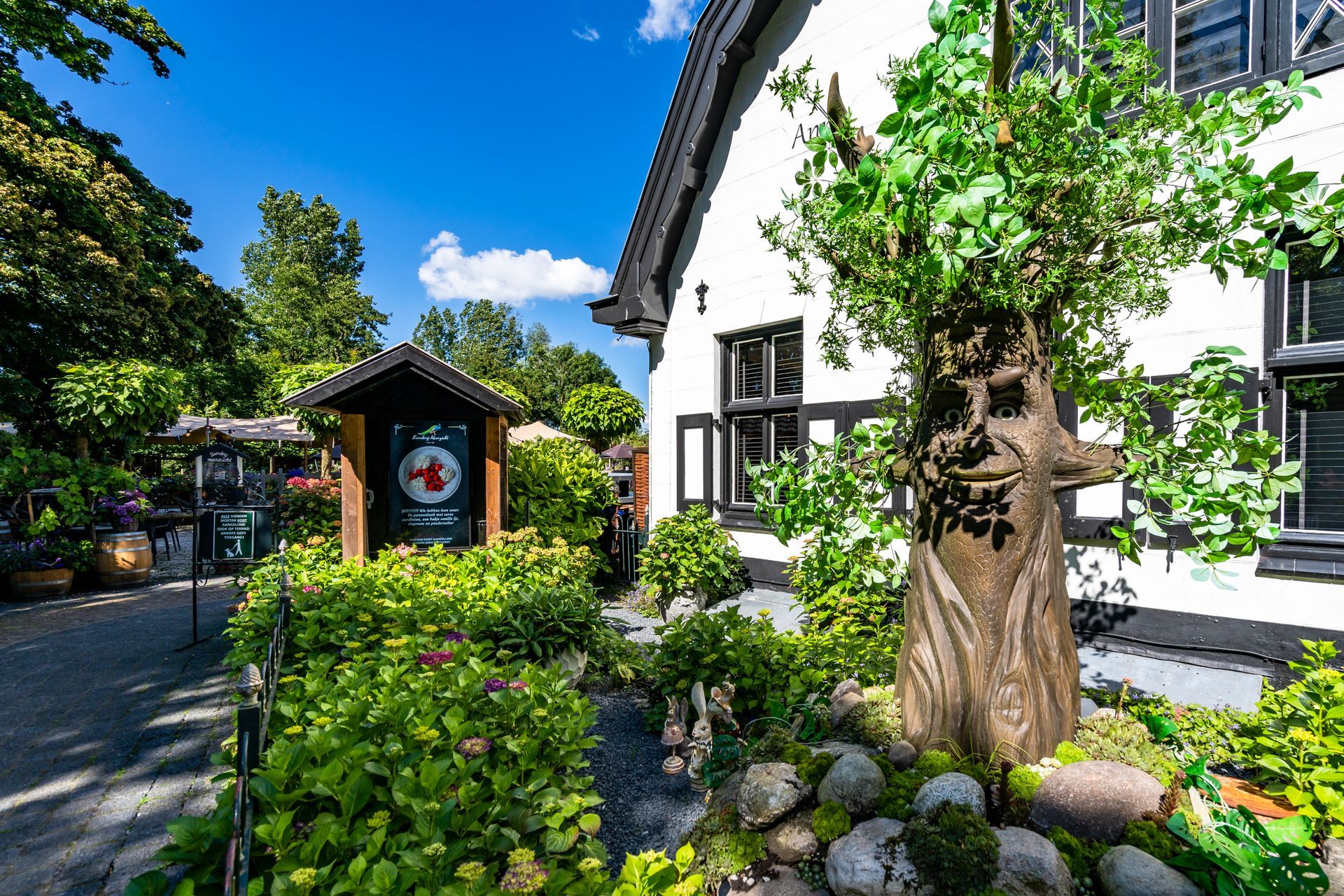 Wit gebouw met zwarte accenten, een uithangbord en een tuin met een grote boom.