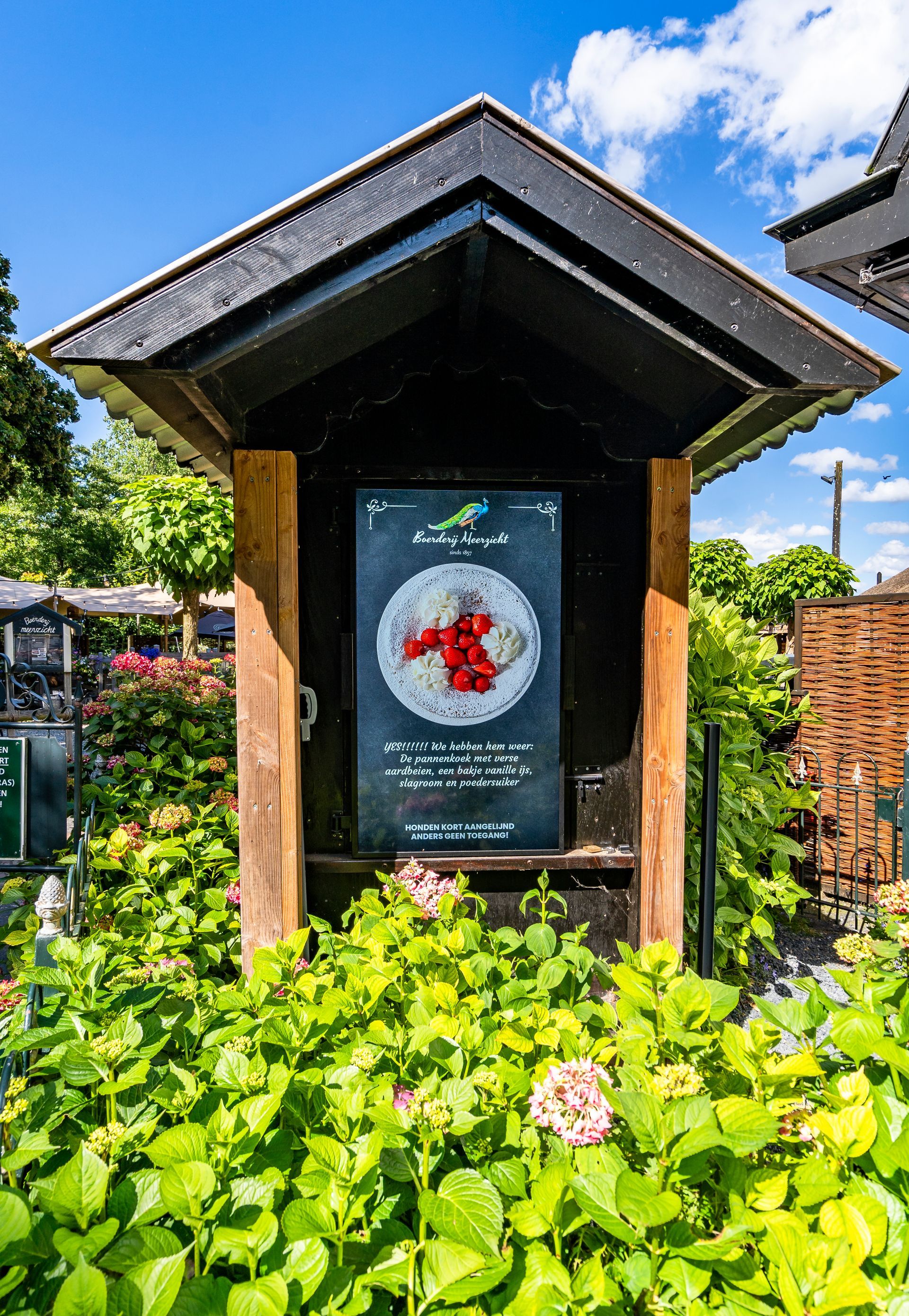 Een bord in een tuin, met een diagram en tekst, onder een zwart dak. Omgeven door groen gebladerte en bloemen.