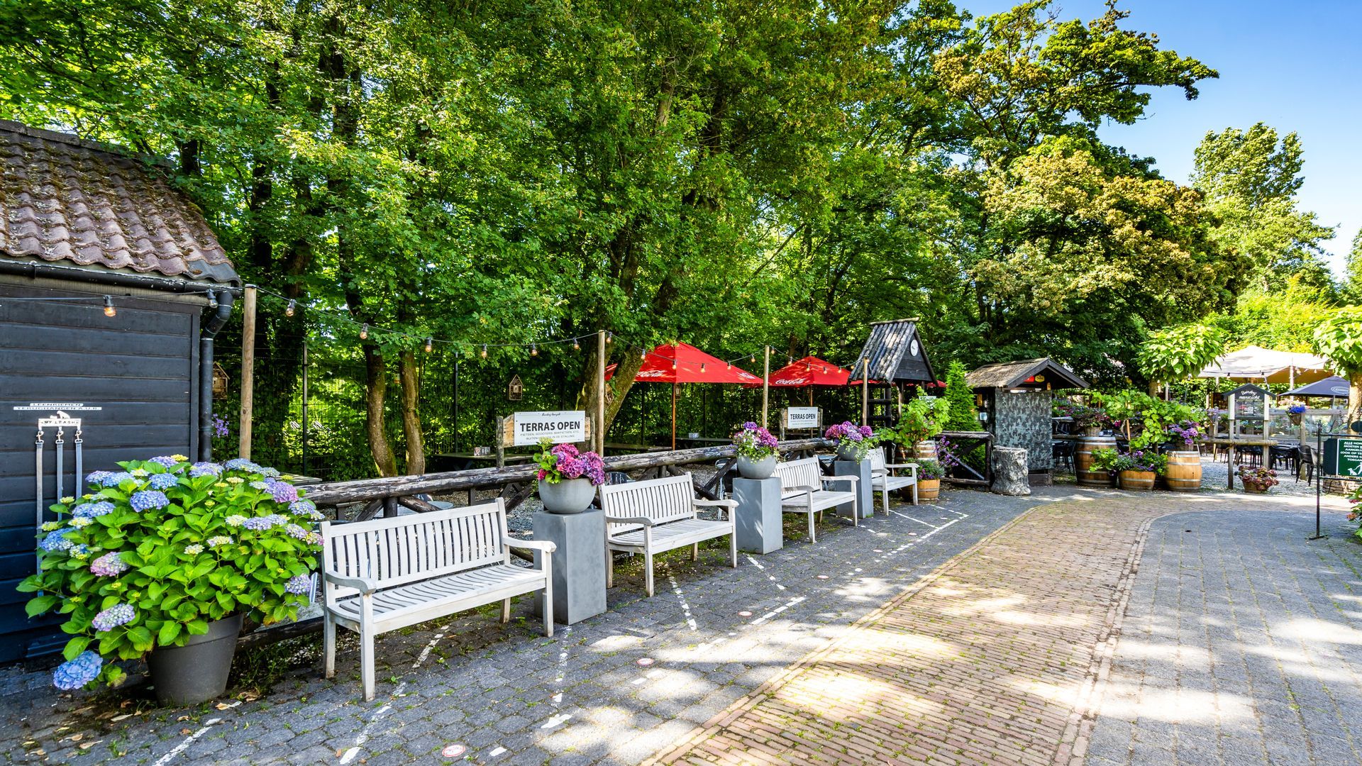 Een zitgedeelte buiten met witte banken, rode parasols en weelderig groen.