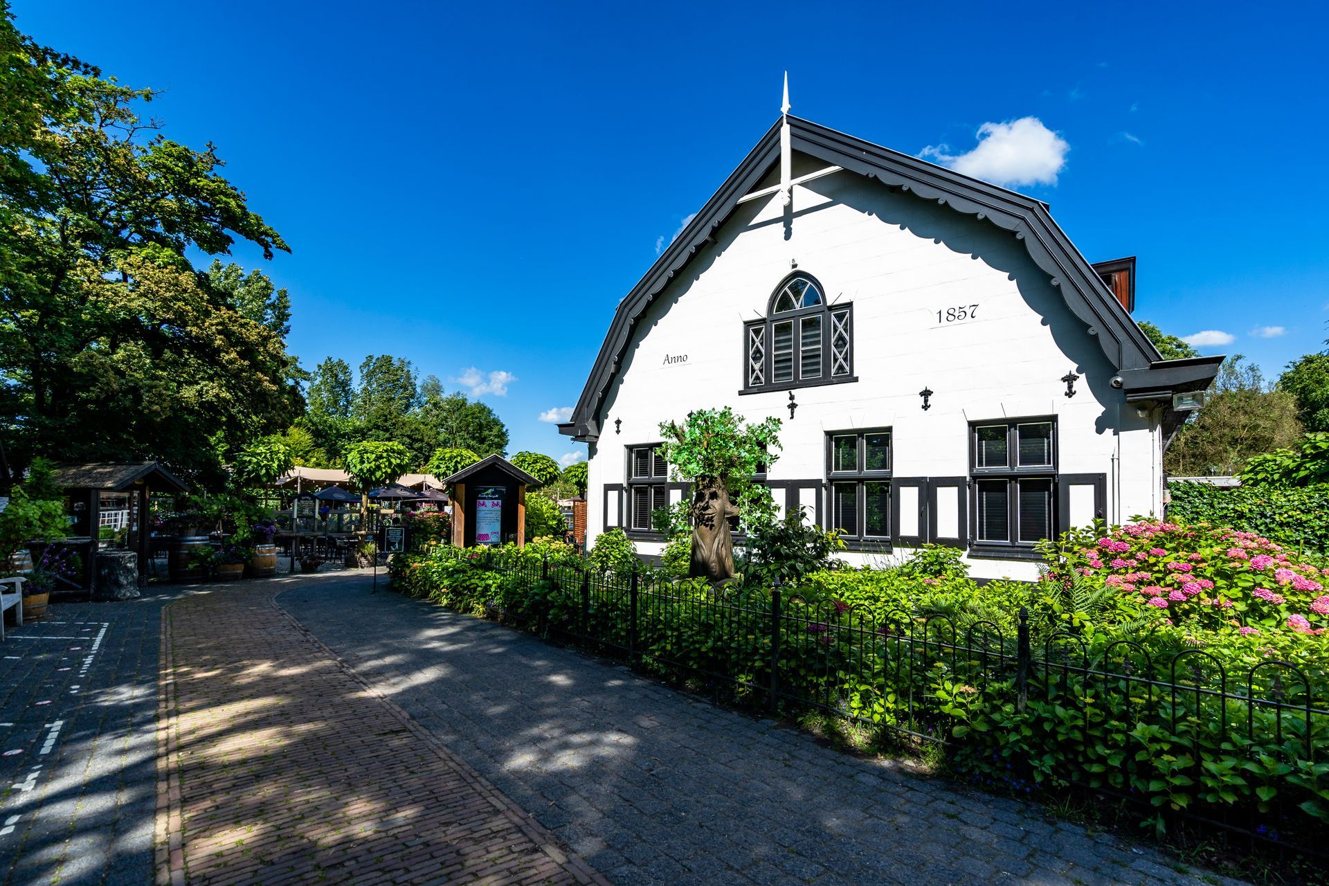 Een wit huis met zwarte accenten en een zadeldak, op een zonnige dag. Een bakstenen pad leidt naar het huis, dat omgeven is door groen.