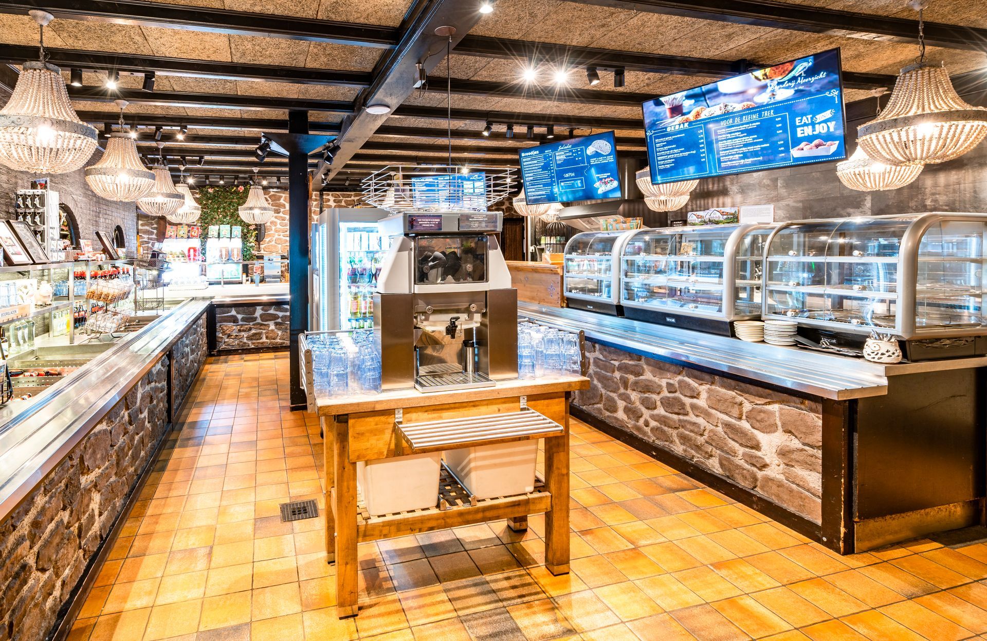 Interieurfoto van een delicatessenwinkel met stenen en houten decoratie, vitrines, tv's en hangende kroonluchters.
