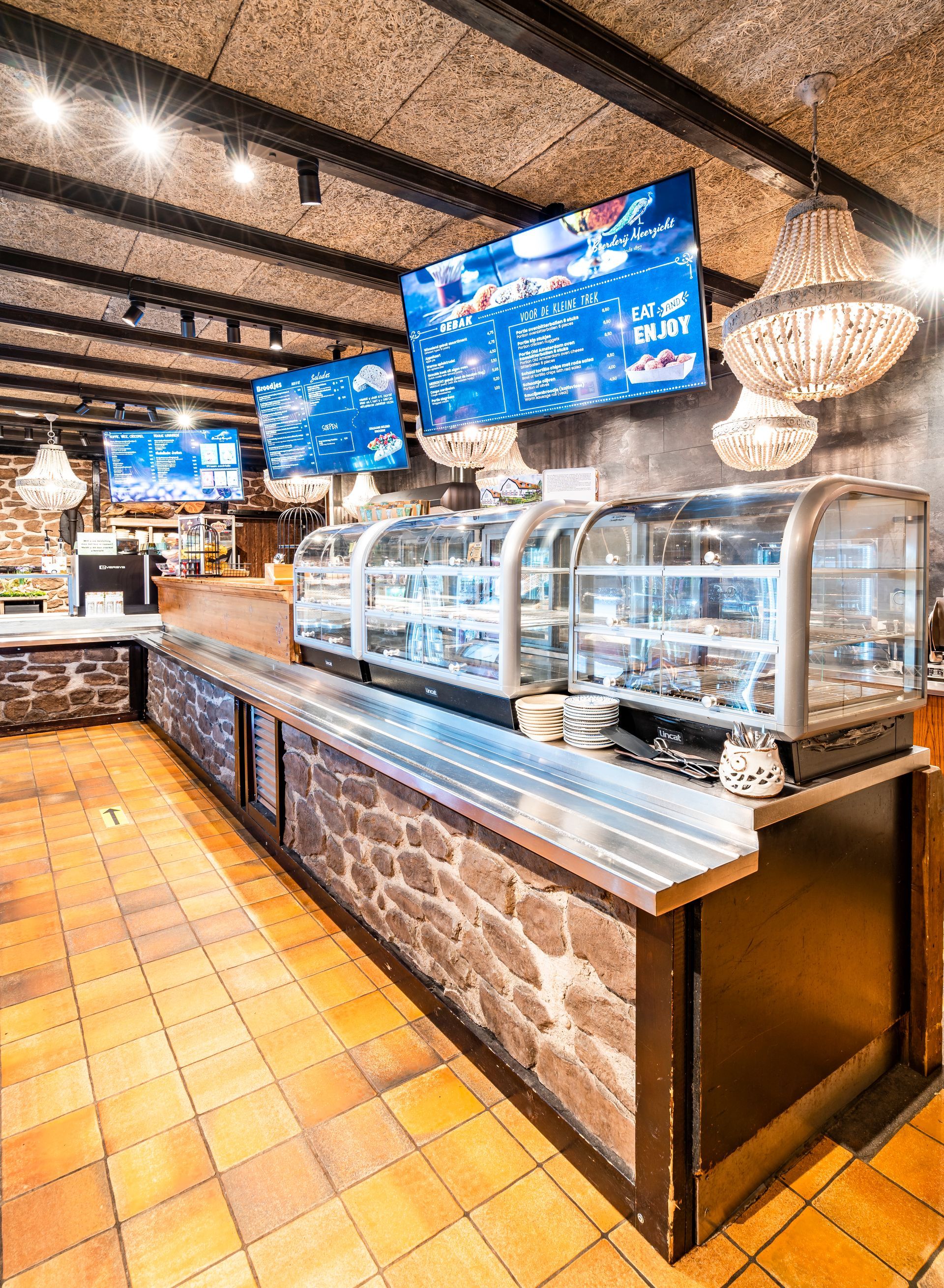 Interieur van een koffiebar met vitrines vol gebak, schermen en decoratieve verlichting.