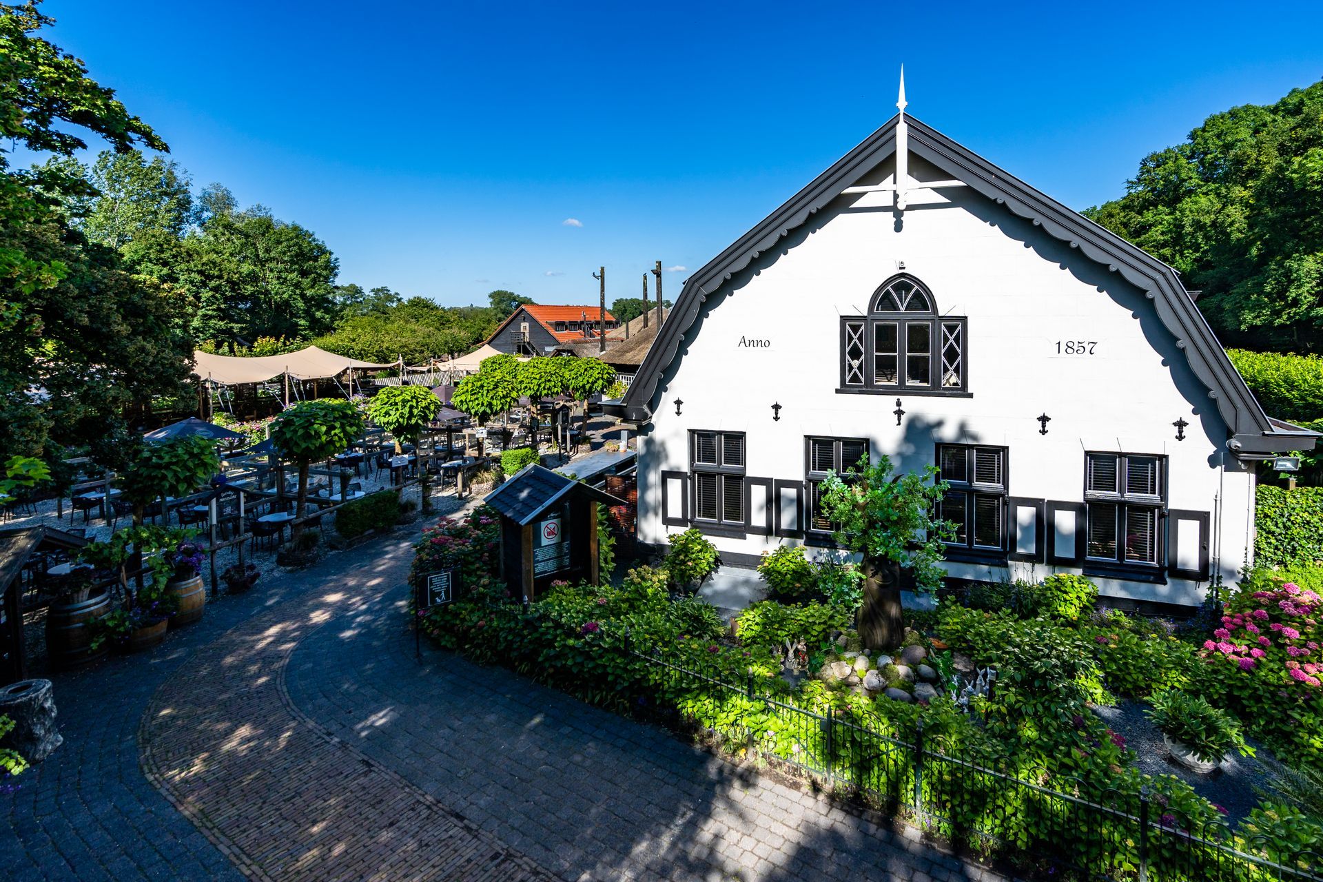 Wit gebouw met zwarte accenten, omgeven door groen en een terras waar op een zonnige dag gegeten kan worden.
