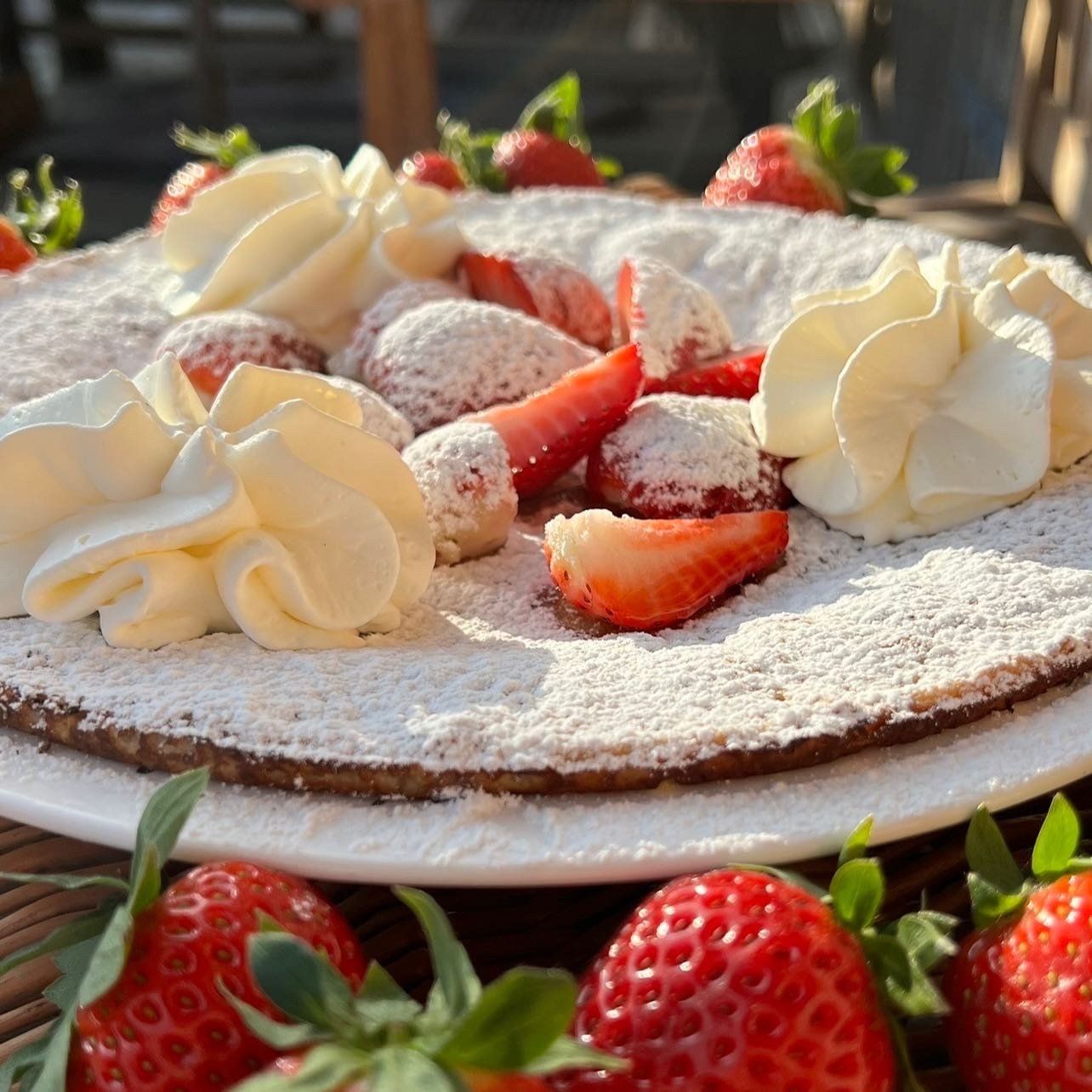 Een pannenkoek, bestrooid met poedersuiker, slagroom en sch schijfjes aardbeien, geserveerd op een wit bord.