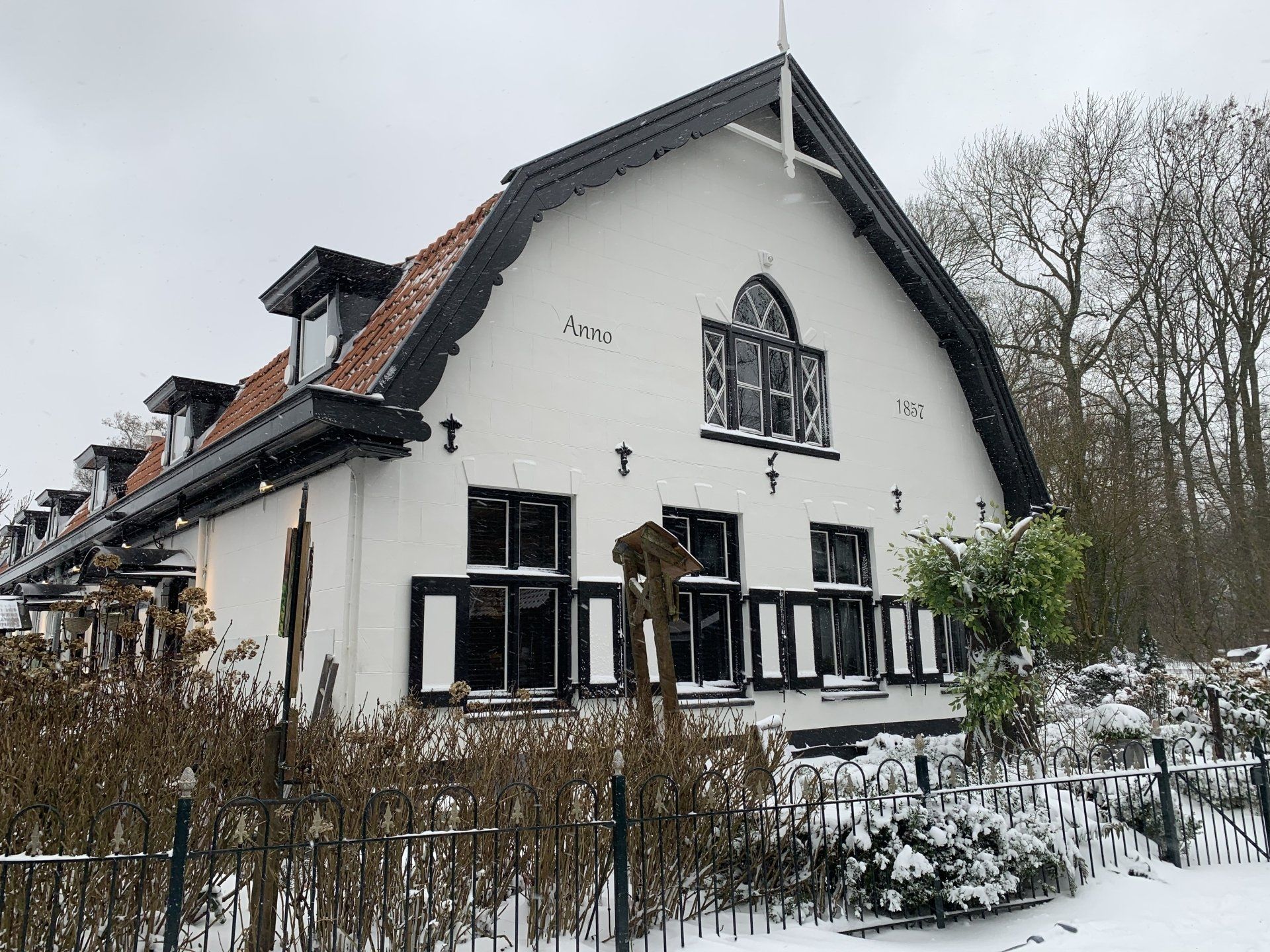 Sneeuwwit huis met zwarte kozijnen en luiken, rood dak, achter een met sneeuw bedekte schutting en struiken.