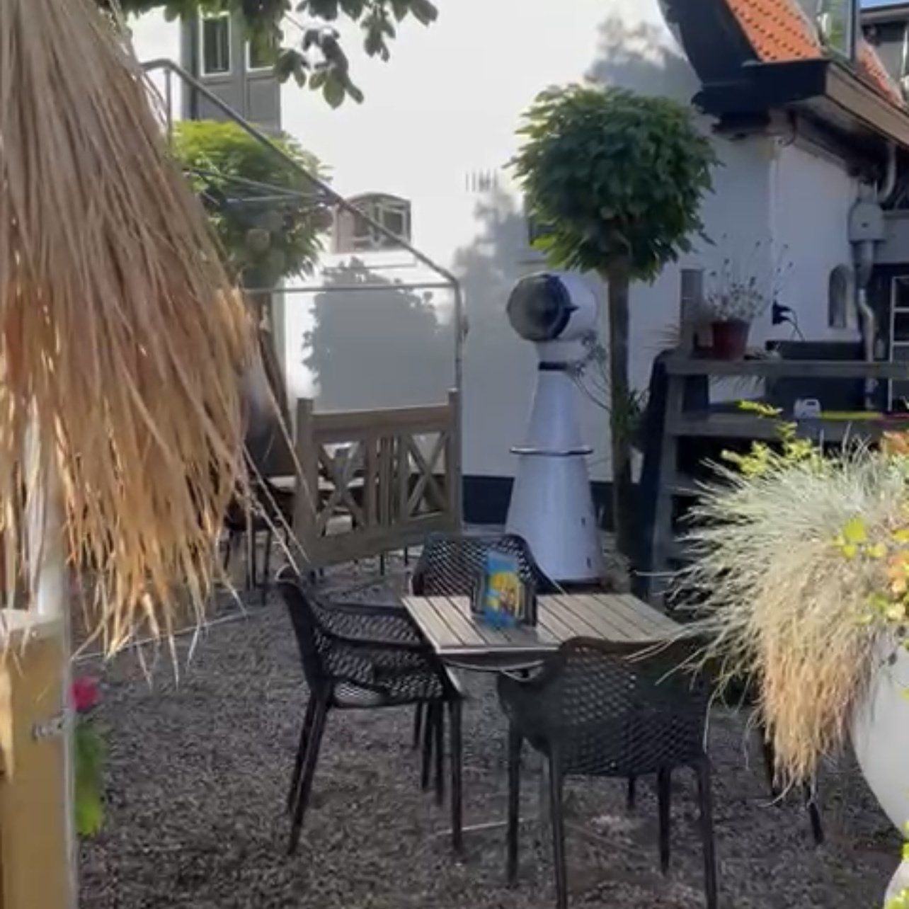 Een terras met tafels en stoelen, een ventilator en groen, tegen de achtergrond van een wit gebouw.