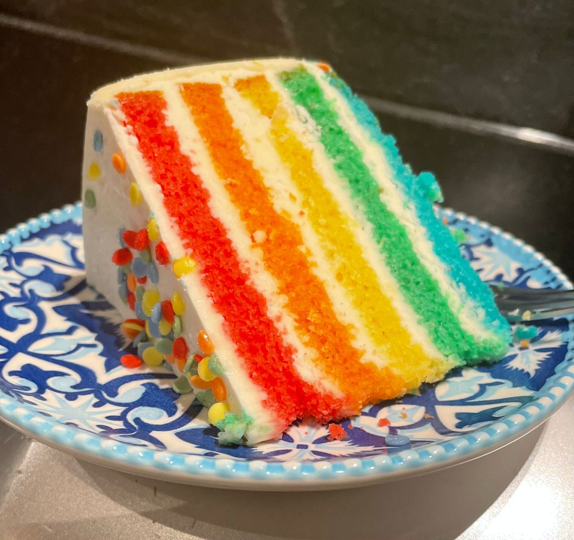 Een stuk regenboogtaart op een blauw-wit bord met hagelslag.
