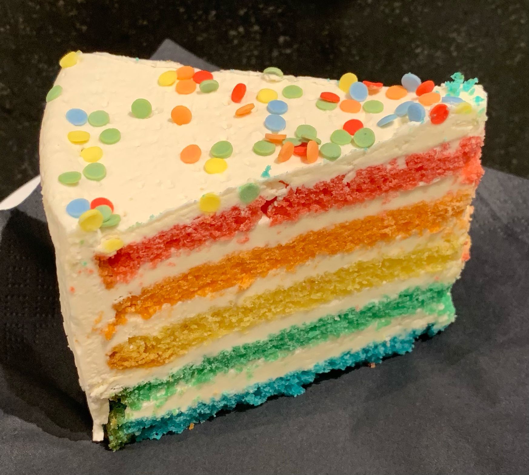 Een plak regenboogtaart met witte glazuur en hagelslag.