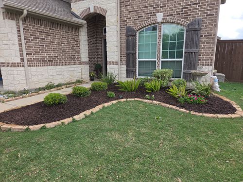 Brick house with flower bed edged with stones, filled with red mulch, and several green bushes.