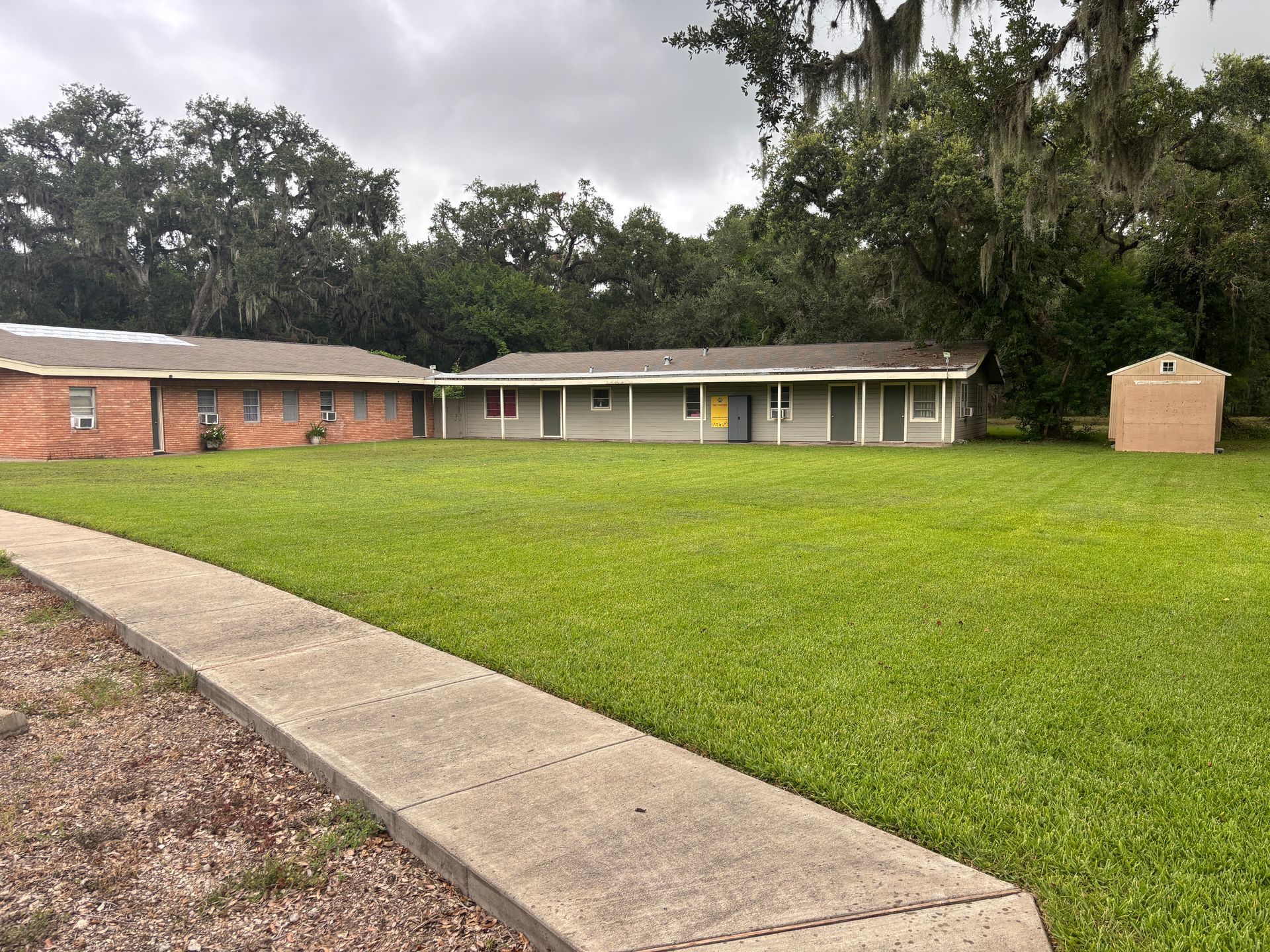 A single-story building with multiple units, green grass lawn, cloudy sky. A sidewalk leads to the buildings.
