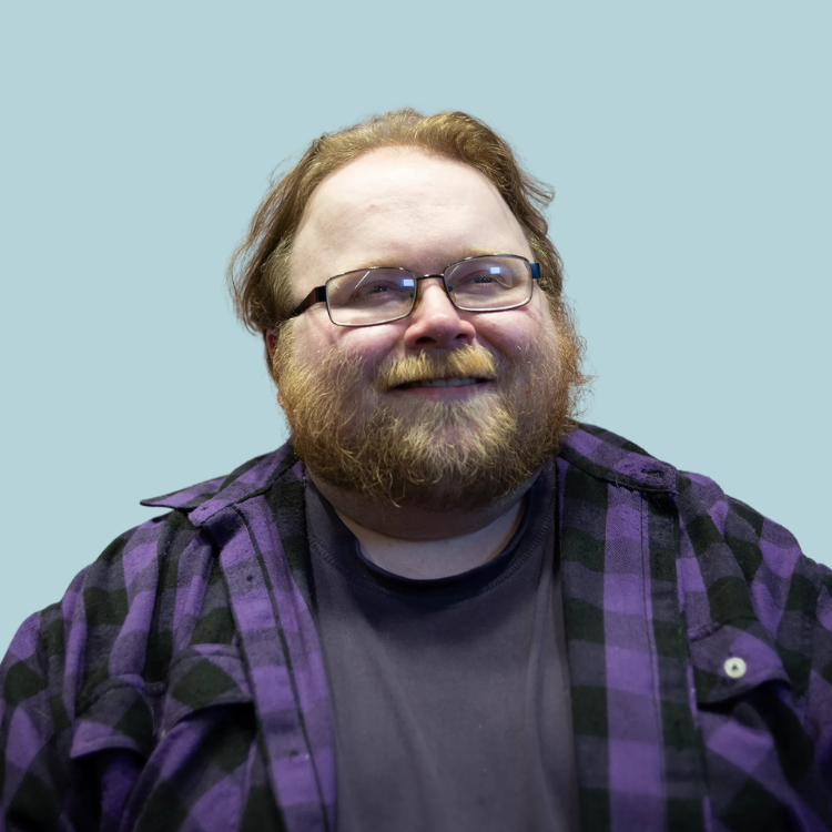 Man with beard and glasses in a purple plaid shirt smiles at the camera. Light blue background.