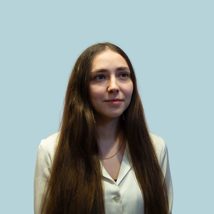 Woman with long brown hair, wearing a cream blazer, looking slightly to the side. Pale blue background.