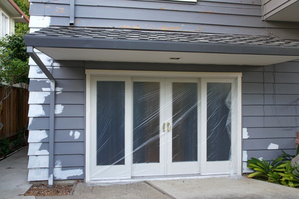 White-framed sliding doors covered in plastic on a gray-sided building with peeling paint.