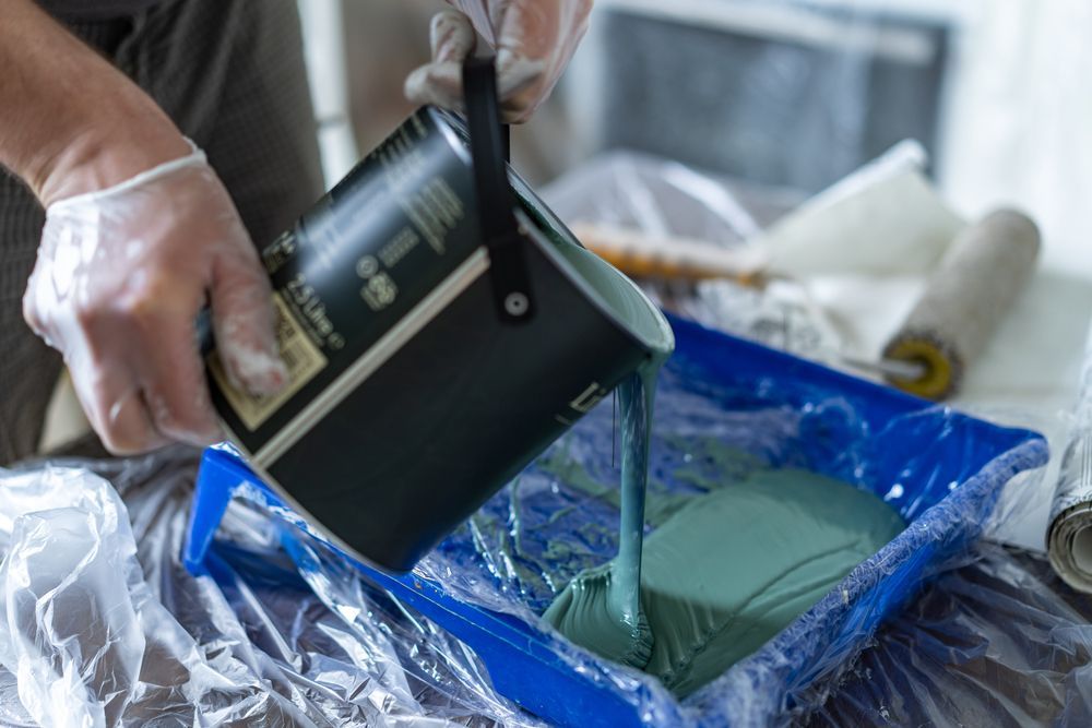 Person pouring teal paint from a can into a blue paint tray lined with plastic, preparing to paint.