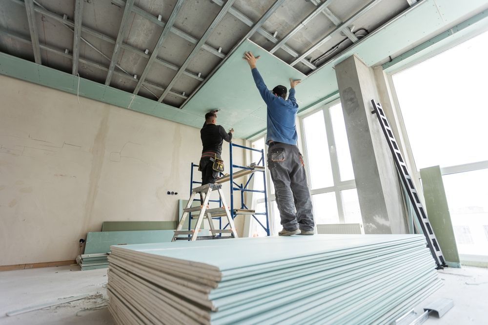 Two workers installing drywall on a ceiling in a room with a large window and stacks of drywall.