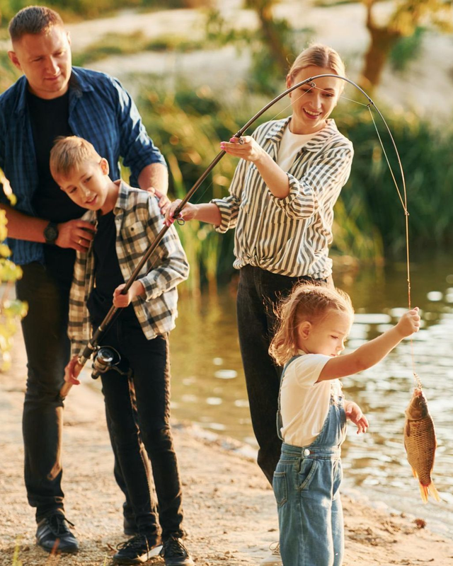 Family fishing by a lakeside, with two adults and two children holding rods and a catch dangling from one line