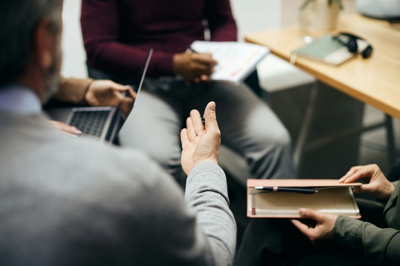 People in a meeting, one speaking while others take notes and use a laptop at a table.