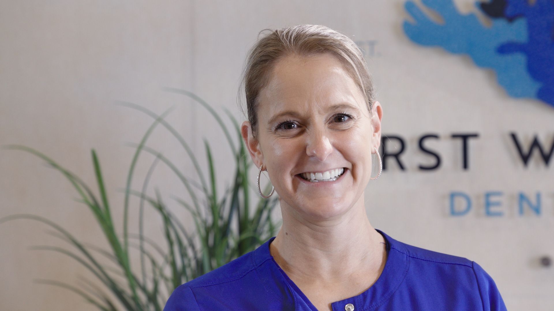 A woman in a blue shirt is smiling in front of a sign.