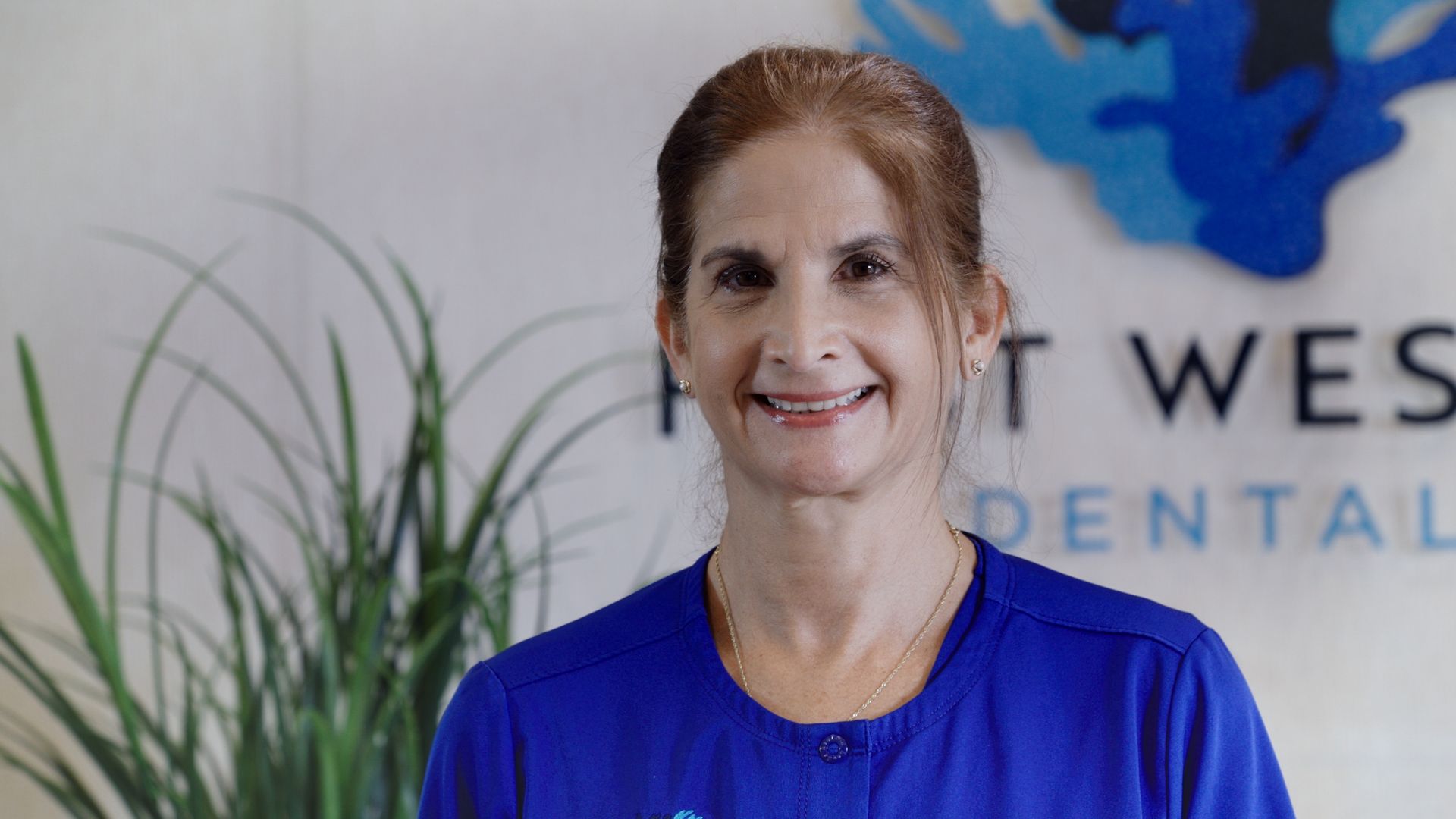 Woman in blue scrubs smiles in front of a dental office logo and plant.