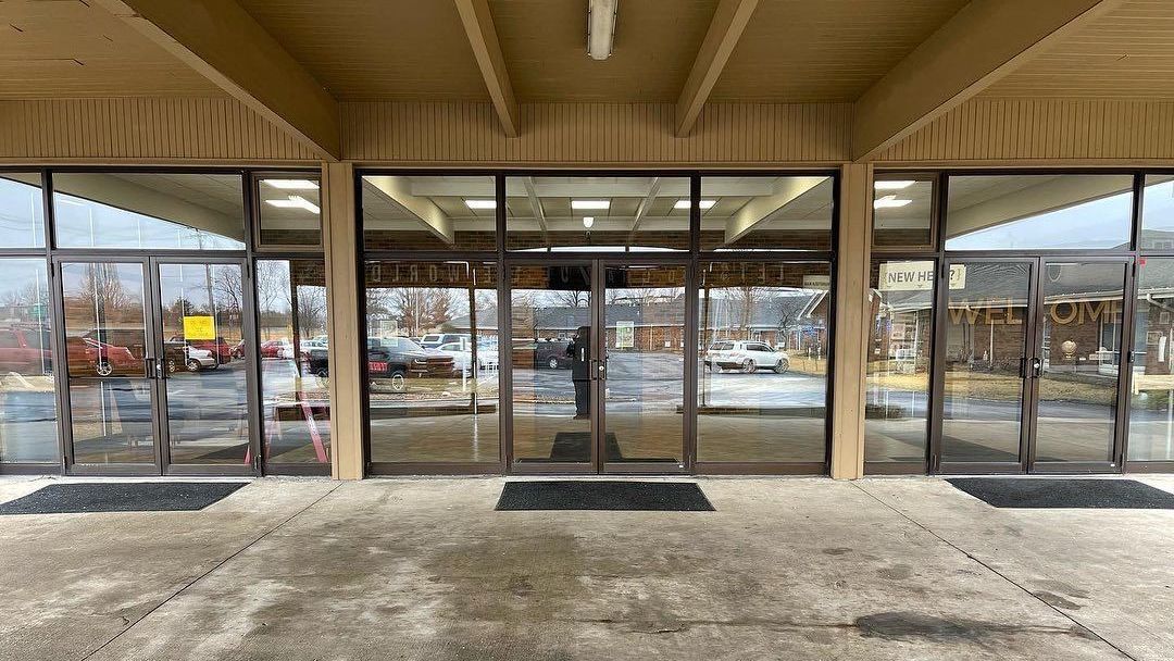 Storefront with glass doors and windows under a covered walkway, reflected parking lot visible outside