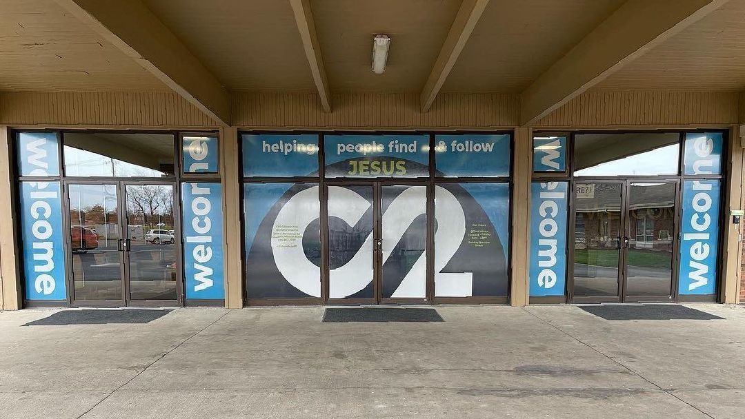 Storefront entrance with blue “welcome” signs and large glass doors under a covered walkway.