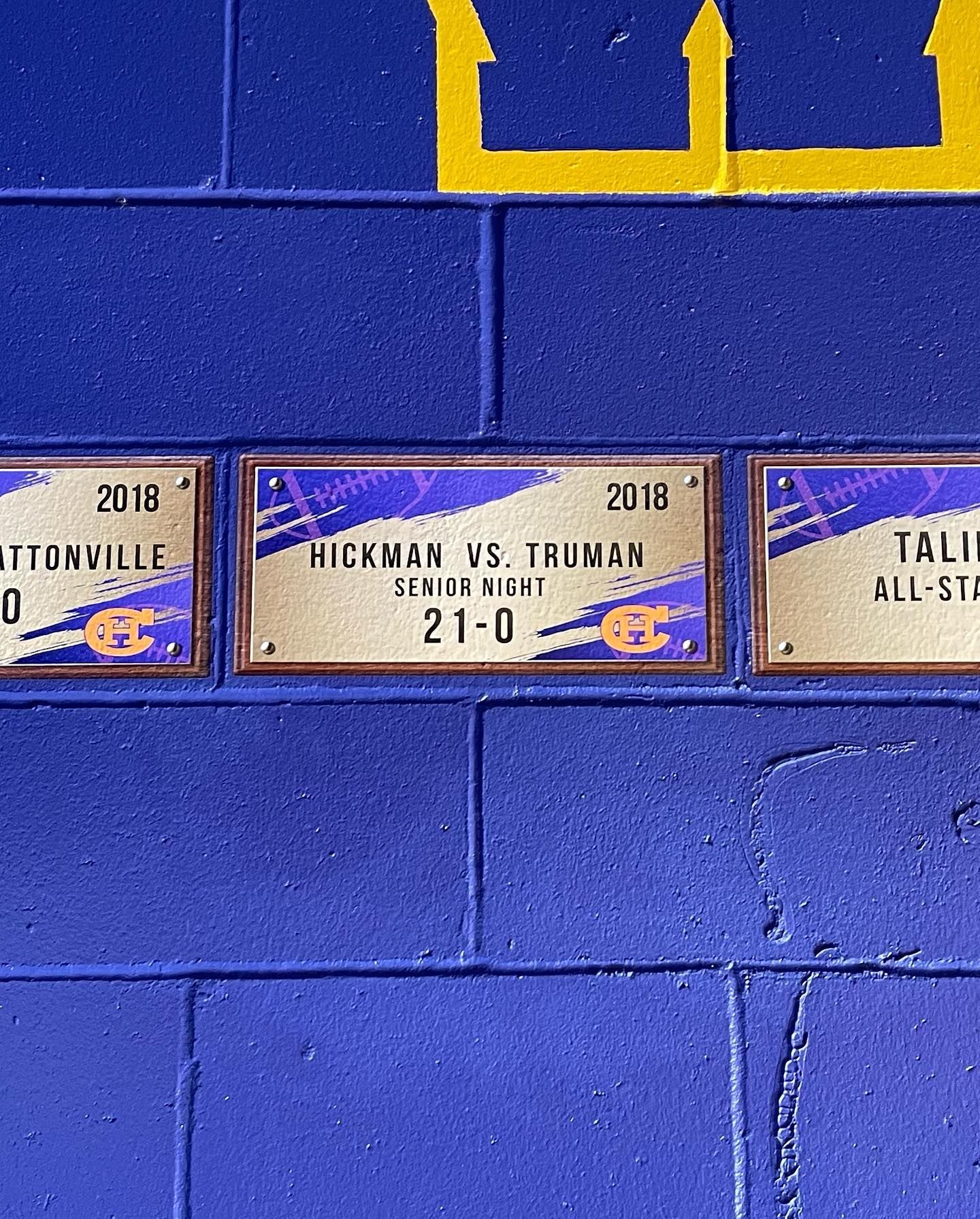 Blue wall with several small framed sports plaques and a yellow crown logo above them.