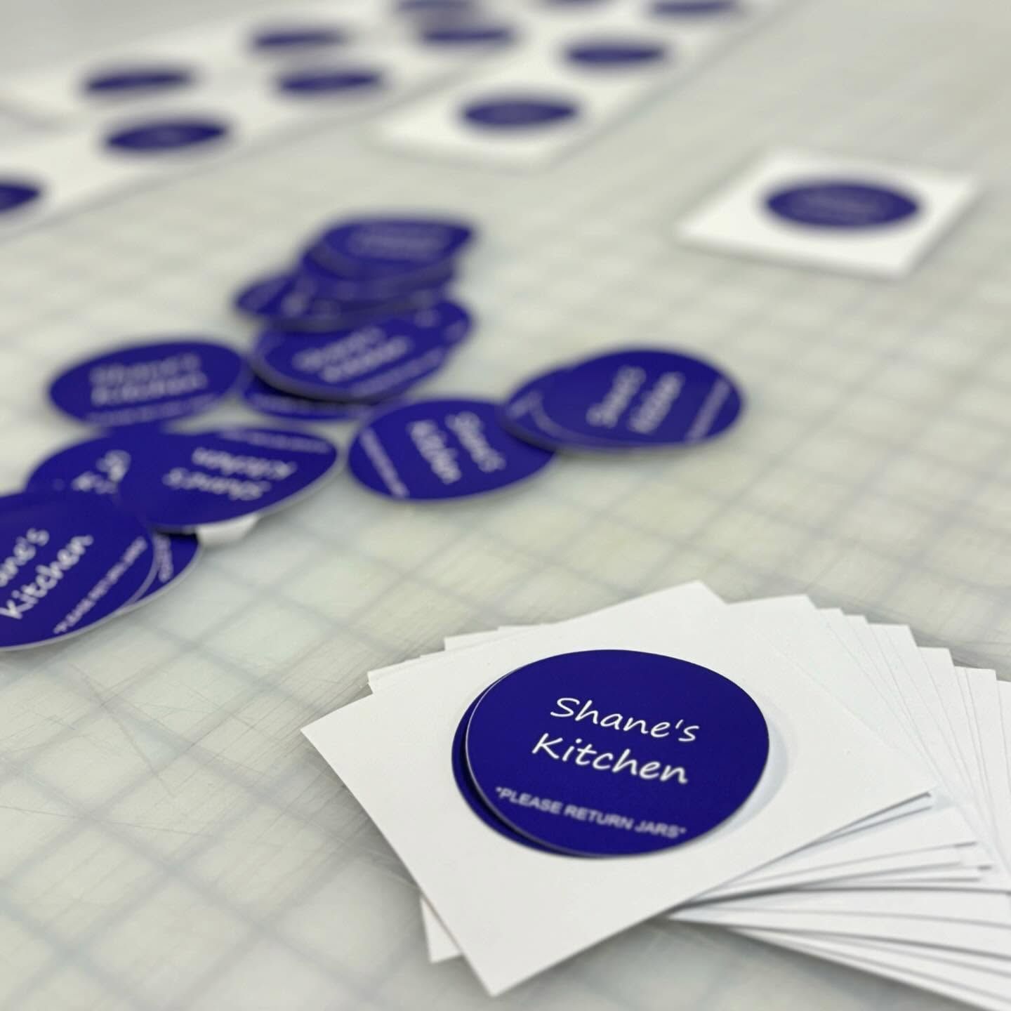 Blue circular logo stickers and stacked business cards on a grid table, reading “Shane’s Kitchen.”