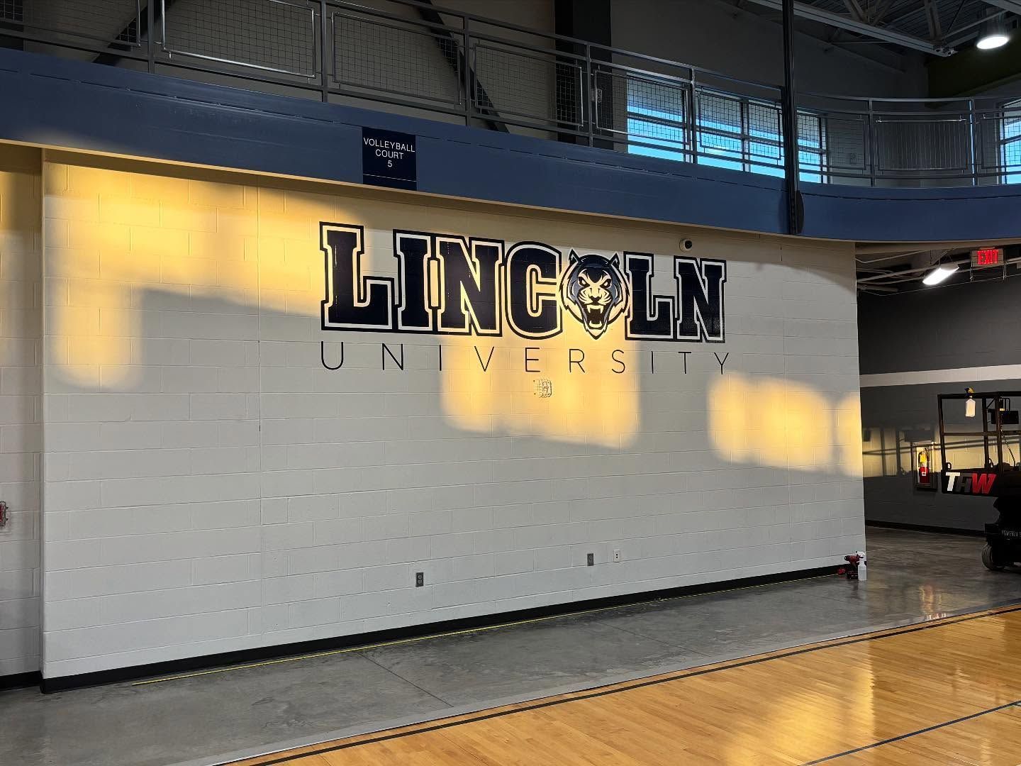 Lincoln University wall sign in a gym, with white tiles and sunlight casting shadows.
