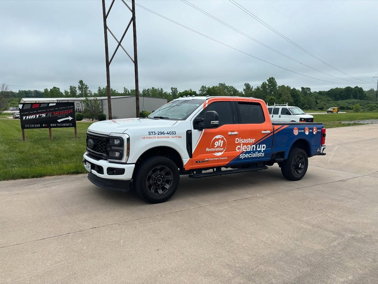 White and orange pickup truck with company branding parked on a roadside near power lines and grass
