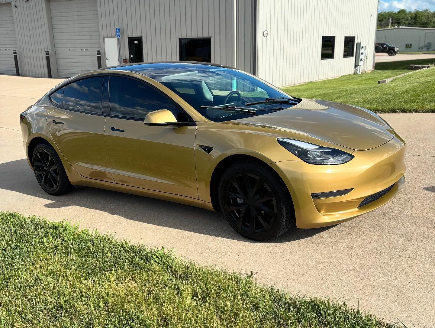 Gold Tesla parked on a sidewalk beside a white industrial building, with black wheels and tinted windows