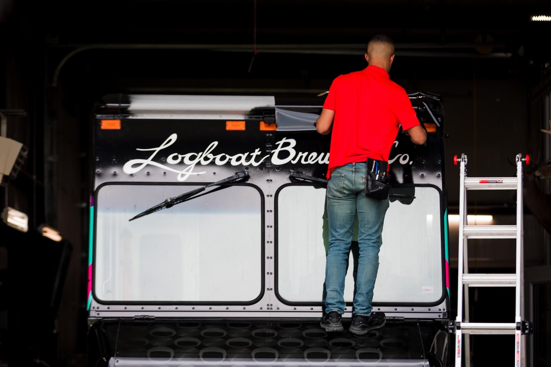 Person in red shirt standing on a truck bed, working near a branded tailgate with a ladder beside it