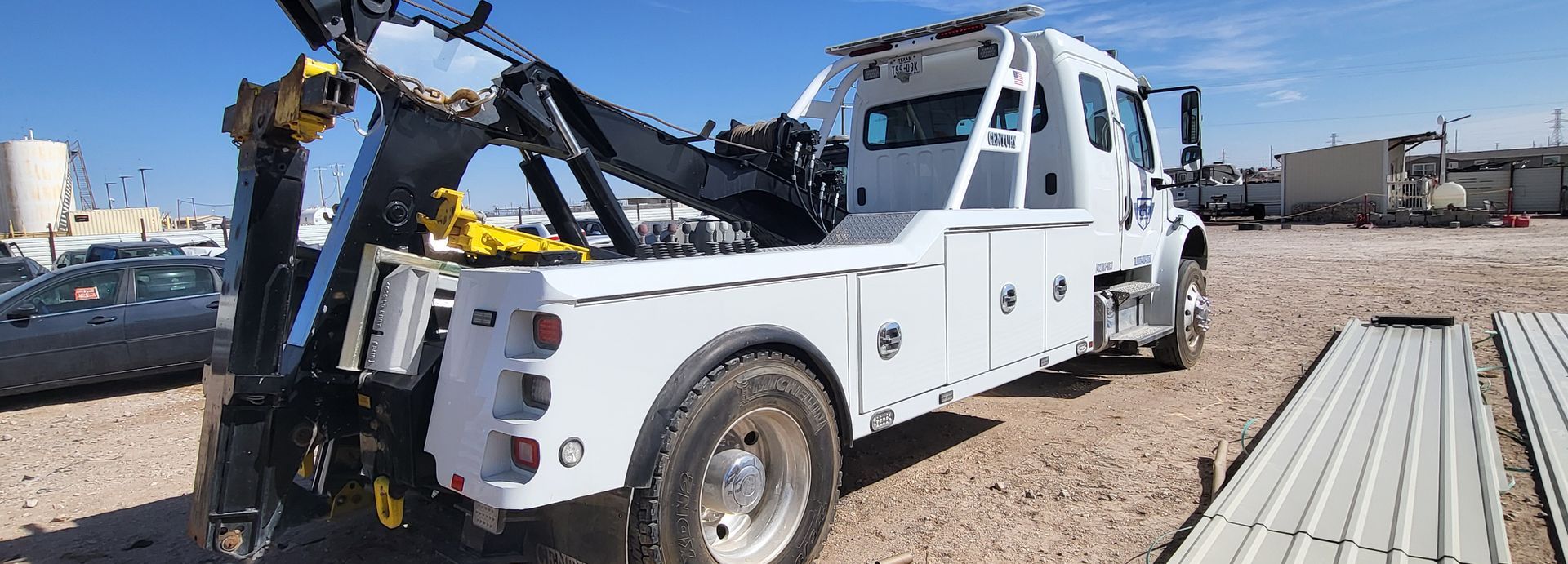 Tow truck lifting a white truck on a gravel lot near stacked metal beams