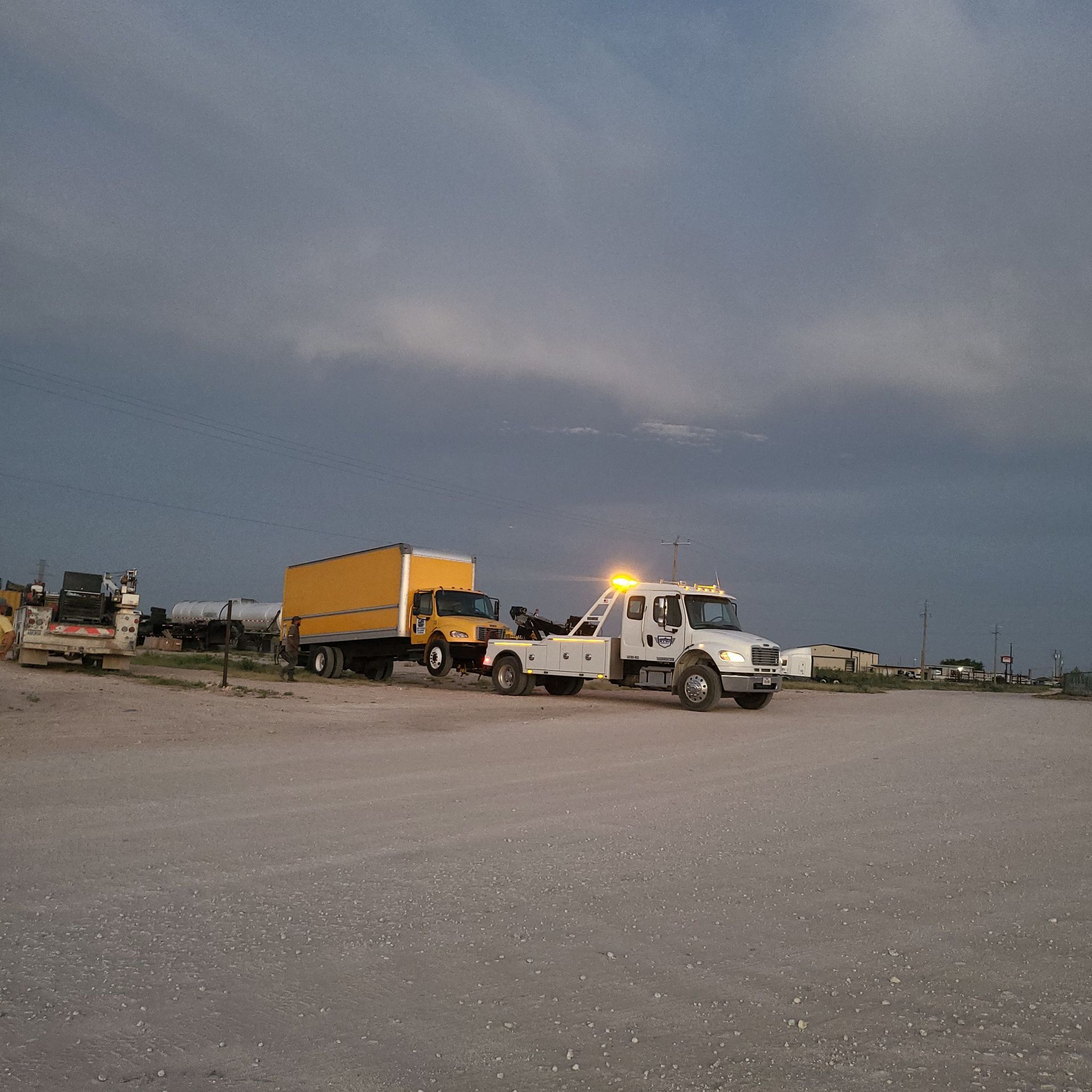Tow truck hauling a yellow dump truck on a gravel lot under a cloudy sky at dusk