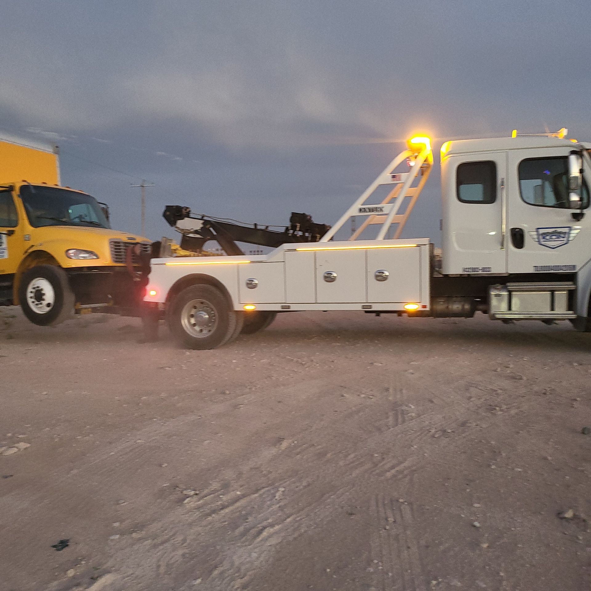 White tow truck with flashing amber lights towing a yellow dump truck at dusk on a gravel lot