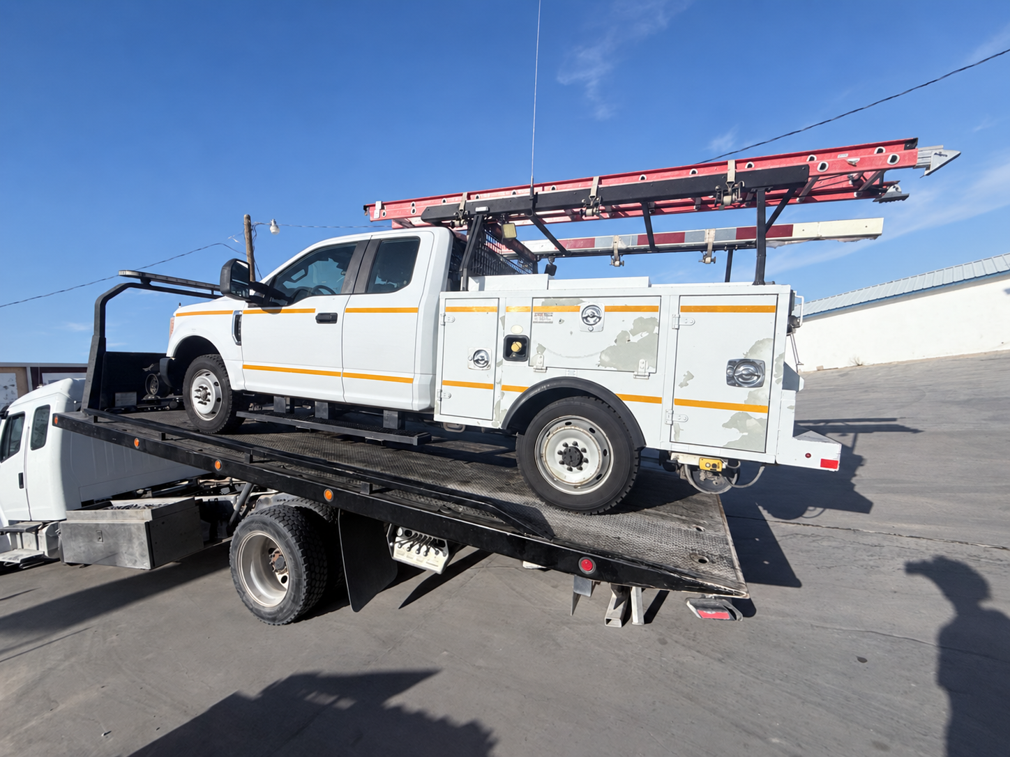 White utility truck with ladder rack loaded on a flatbed tow truck in a parking lot
