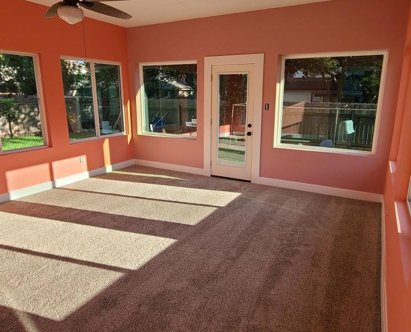 Empty sunroom with salmon walls, tan carpet, and large windows and glass door letting in sunlight.