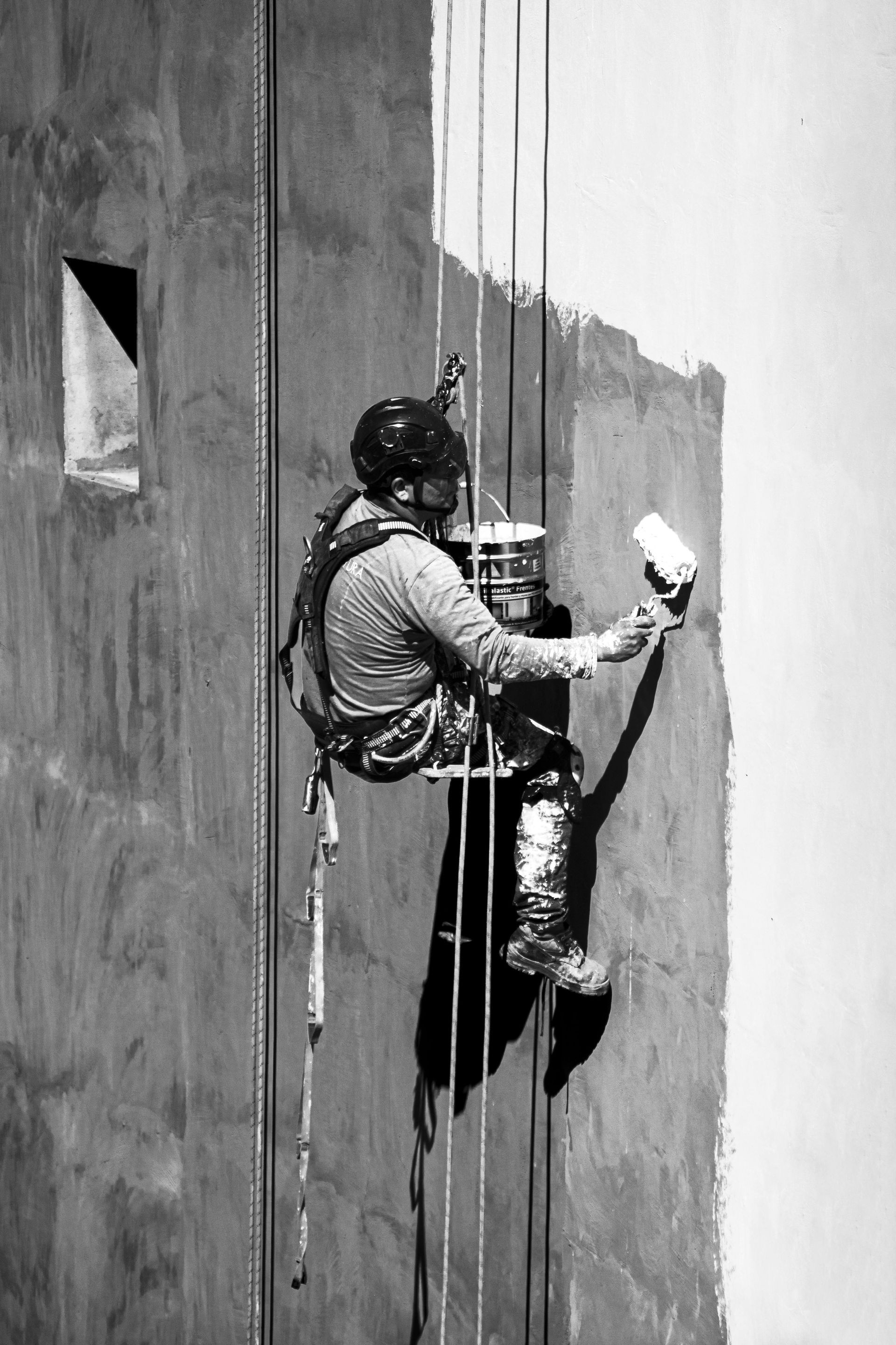 Worker rappels down a tall wall, painting it with a roller and bucket.