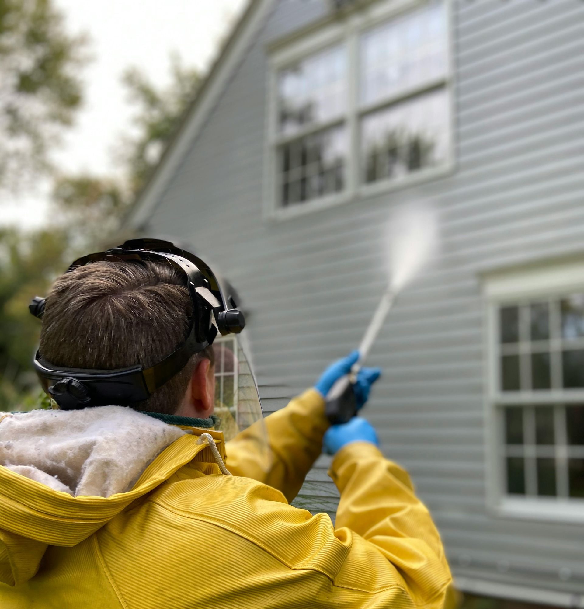 Worker in yellow jacket pressure-washing a gray house exterior with a hose